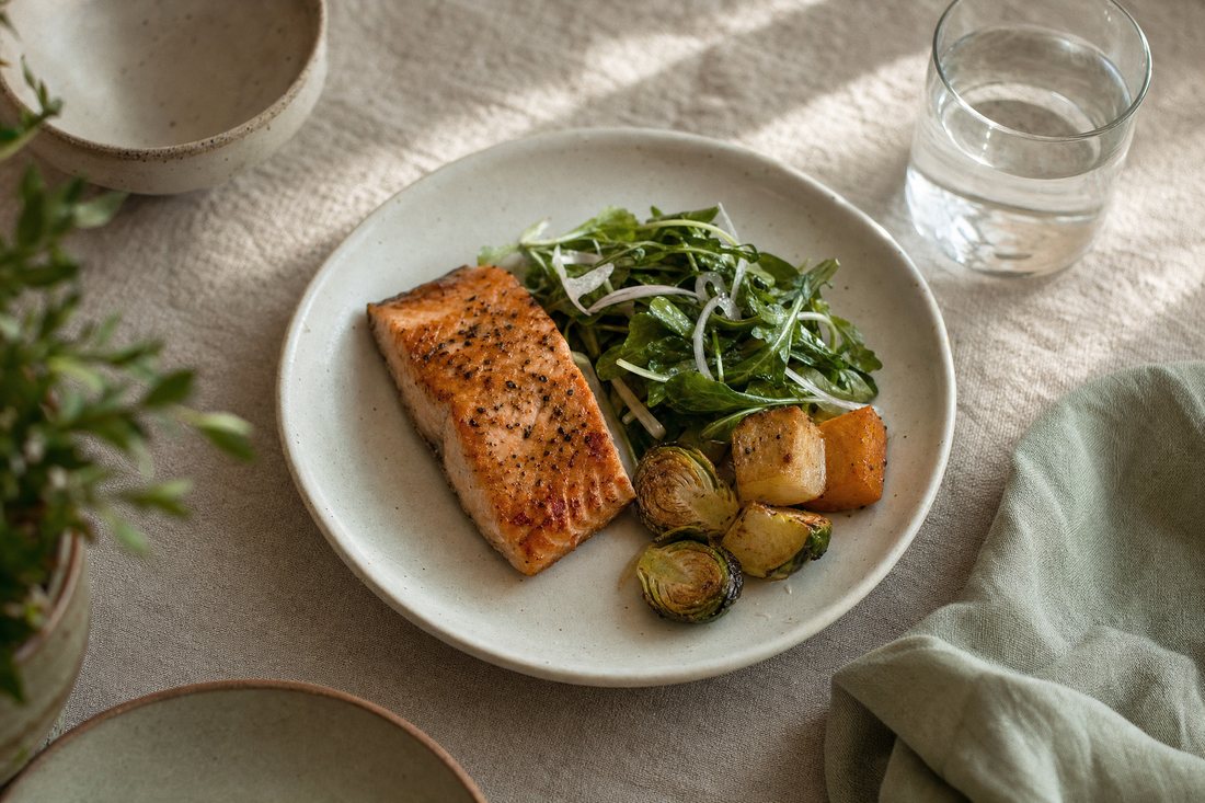 A modest plate of salmon, rocket salad and roasted vegetables beside a glass of water on a linen tablecloth.